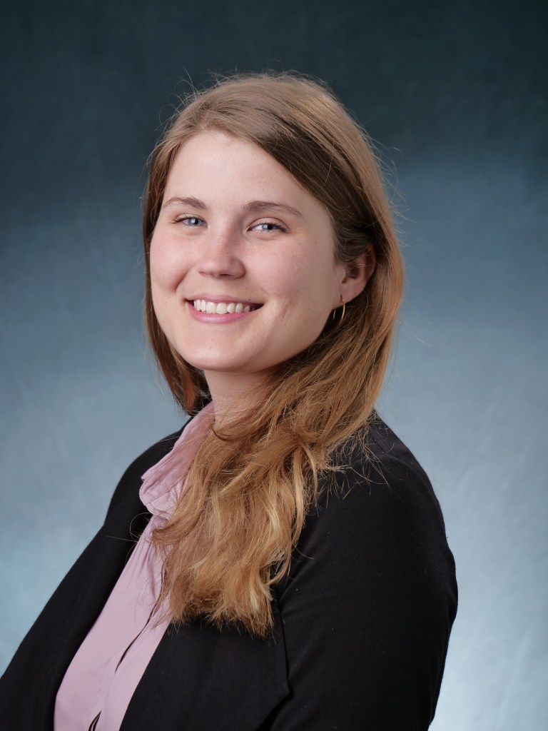 A headshot of Kara with a grey background.  She is a white female with long light brown hair.  Her hair is parted to the side and she is smiling.  She is wearing a light purple shirt with a black jacket and hoop earrings.